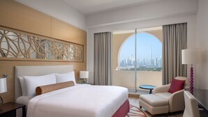 Habitación de hotel con una cama grande, un escritorio, una silla y vistas a un horizonte urbano a través de un ventanal.