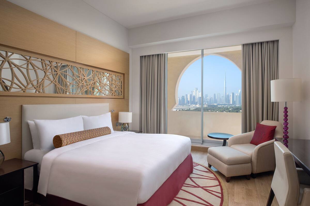 A hotel room with a large bed, a desk, a chair, and a view of a city skyline through a large window.