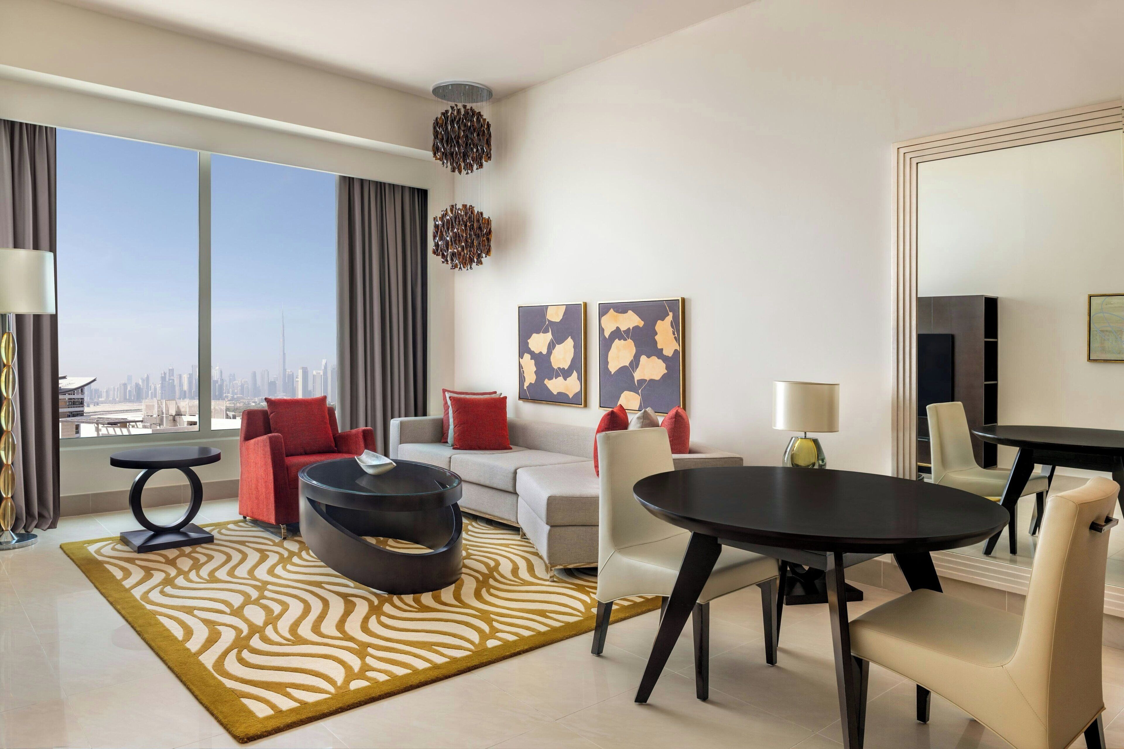 A modern hotel room with a large window offering a city view, a sofa, a dining table, and a black circular ottoman.