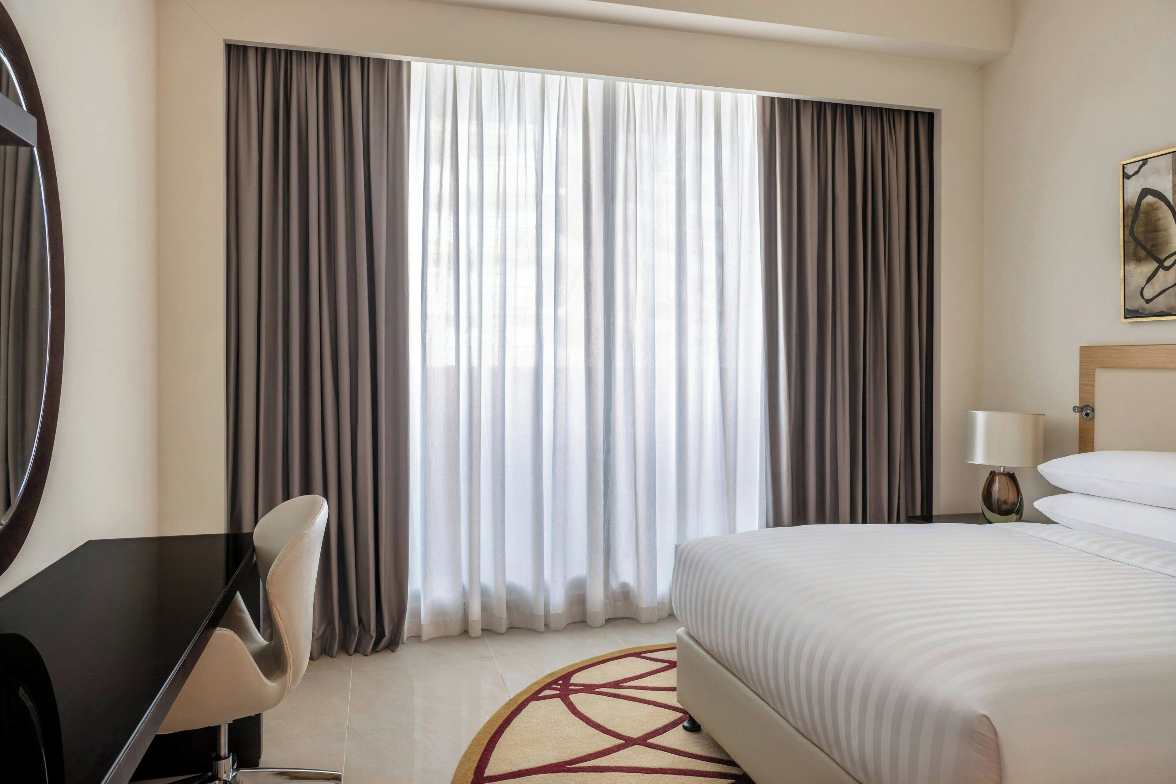 Ein Hotelzimmer mit einem Bett, einem Schreibtisch und einem Stuhl. Es gibt ein Fenster mit Vorhängen, eine Lampe und ein Bild an der Wand.