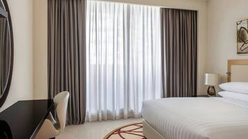 Ein Hotelzimmer mit einem Bett, einem Schreibtisch und einem Stuhl. Es gibt ein Fenster mit Vorhängen, eine Lampe und ein Bild an der Wand.