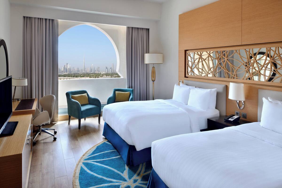 A hotel room with two beds, a desk, a chair, and a view of a cityscape.
