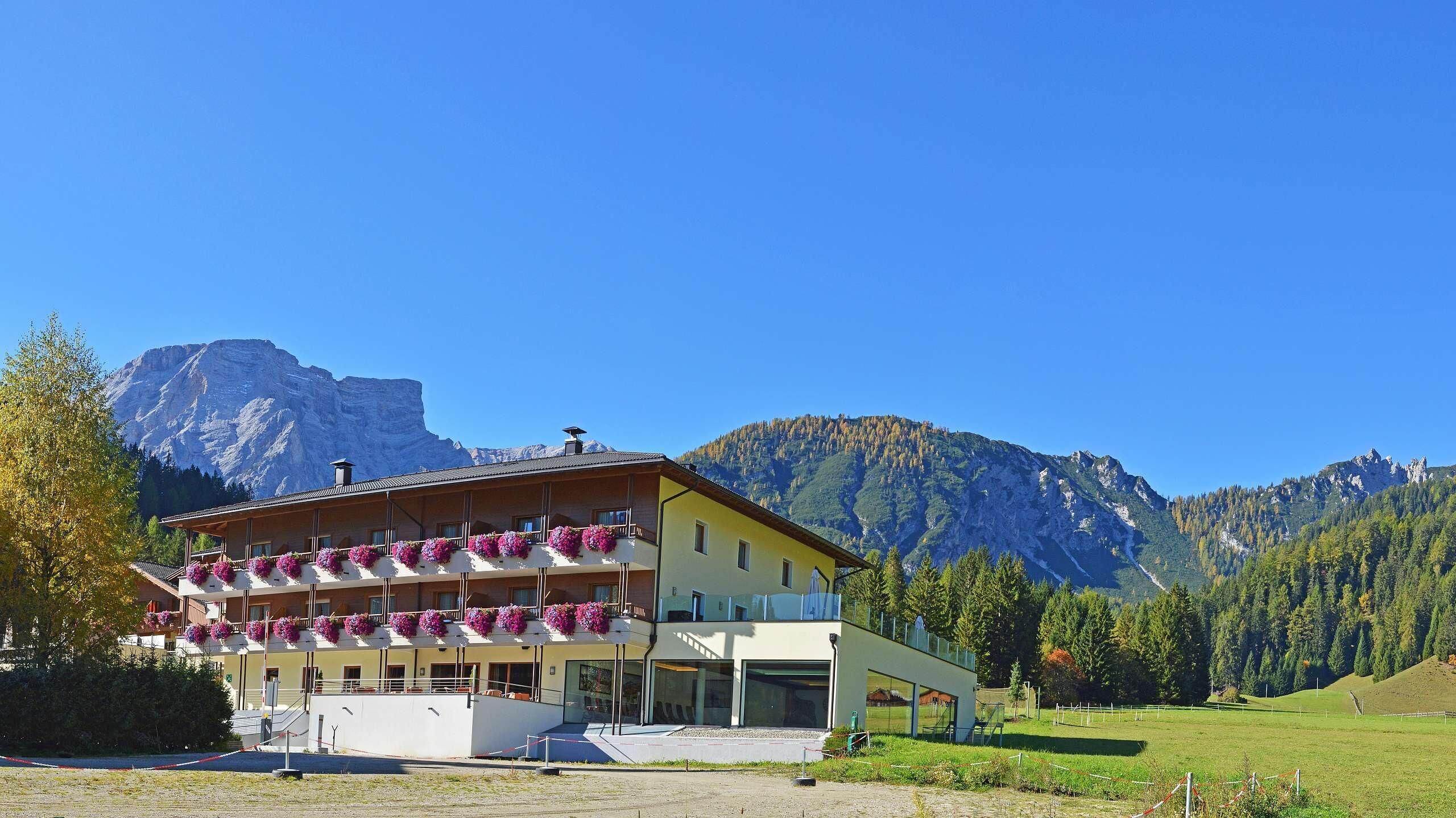 Ein Hotel mit mehreren Blumen geschmückten Balkonen vor einer Bergkulisse und blauem Himmel.