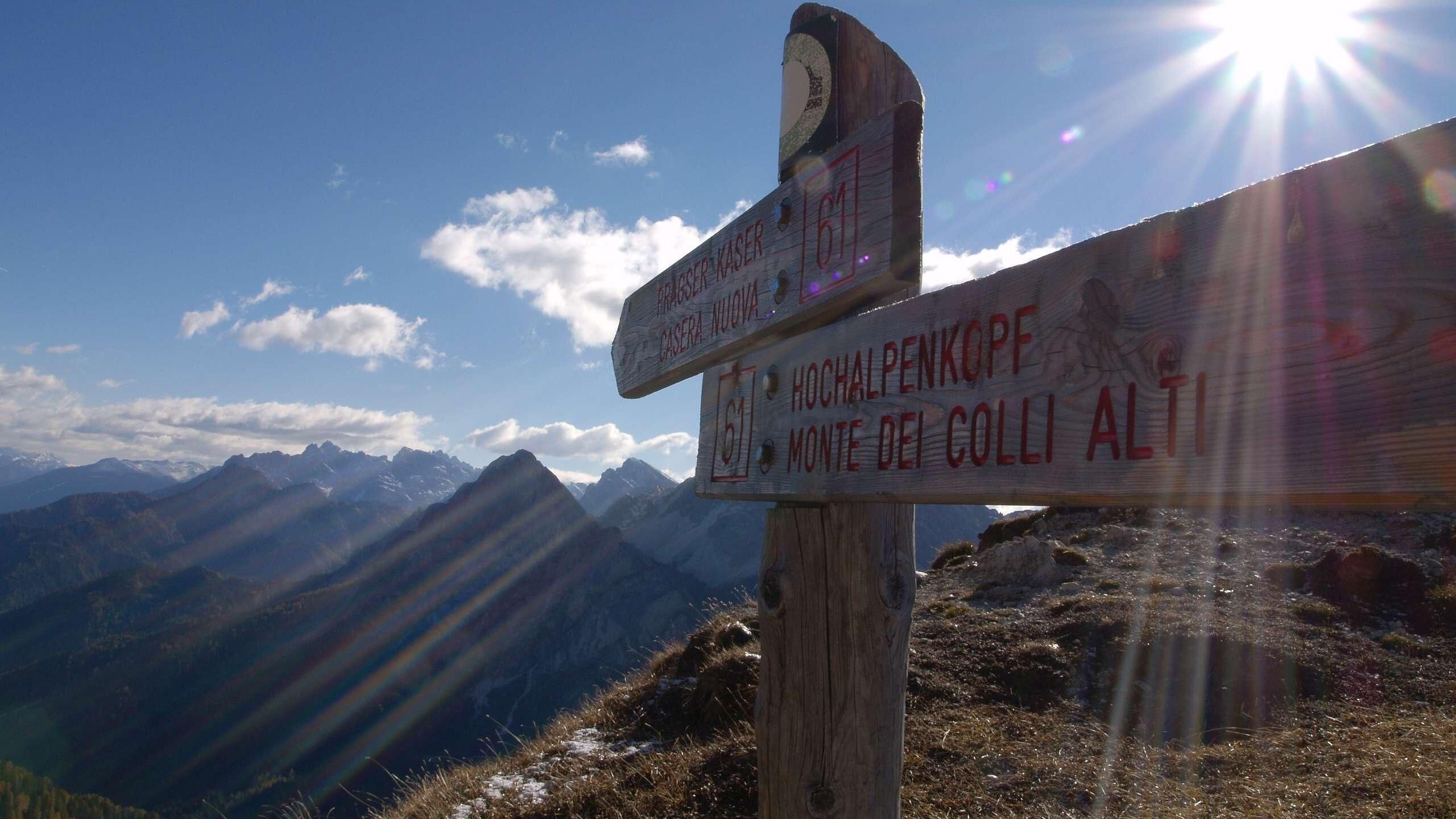 A wooden signpost with directional information for hikers, set against a mountainous backdrop with a clear sky.