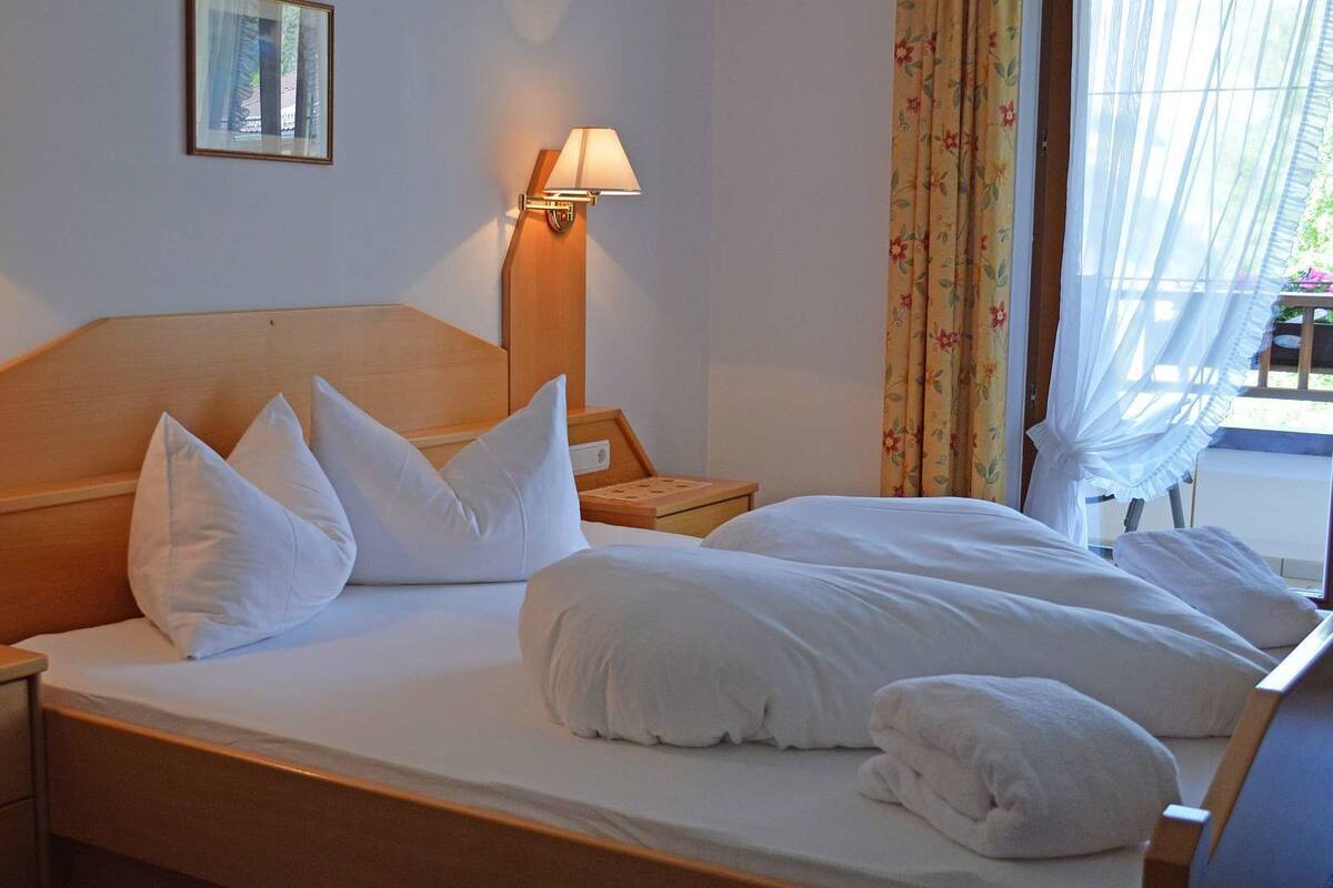 A hotel room with a wooden headboard, a bed with white linens and pillows, two bedside lamps, a nightstand with a telephone, and a window with floral curtains.