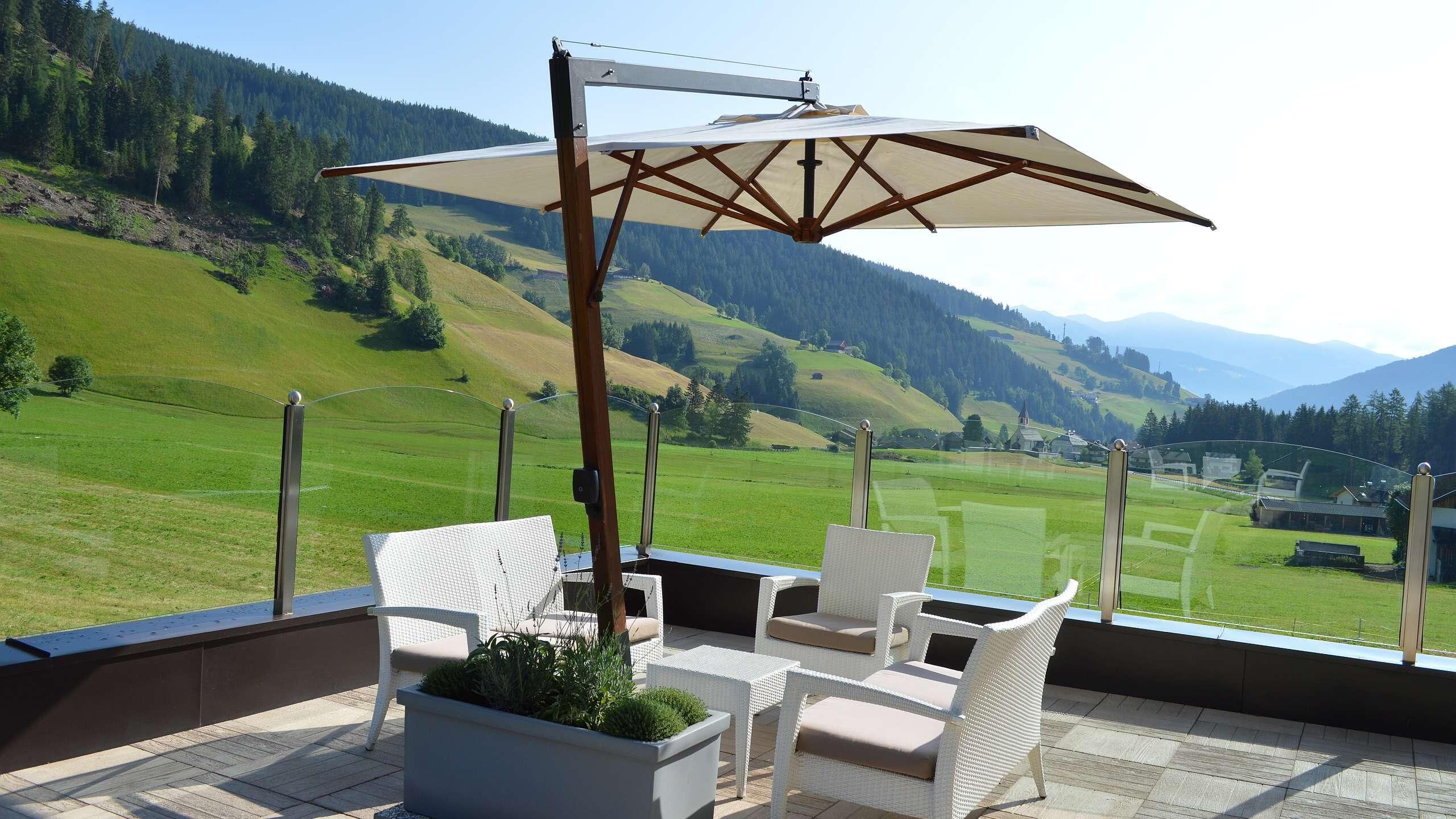 A balcony with white wicker furniture, a large umbrella, and a view of a green valley and mountains.