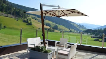 A balcony with white wicker furniture, a large umbrella, and a view of a green valley and mountains.