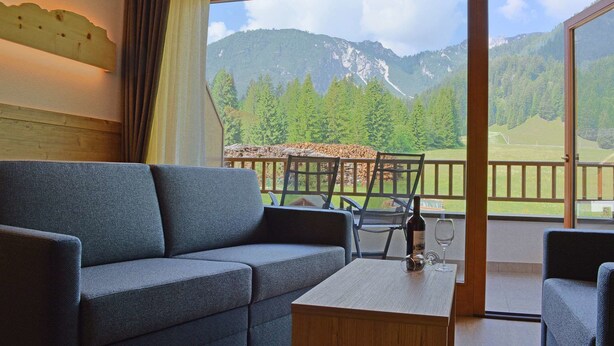 Una habitación con un sofá gris, una mesa de centro de madera y una silla con estructura metálica. Una ventana muestra un paisaje montañoso con árboles y una terraza de madera.