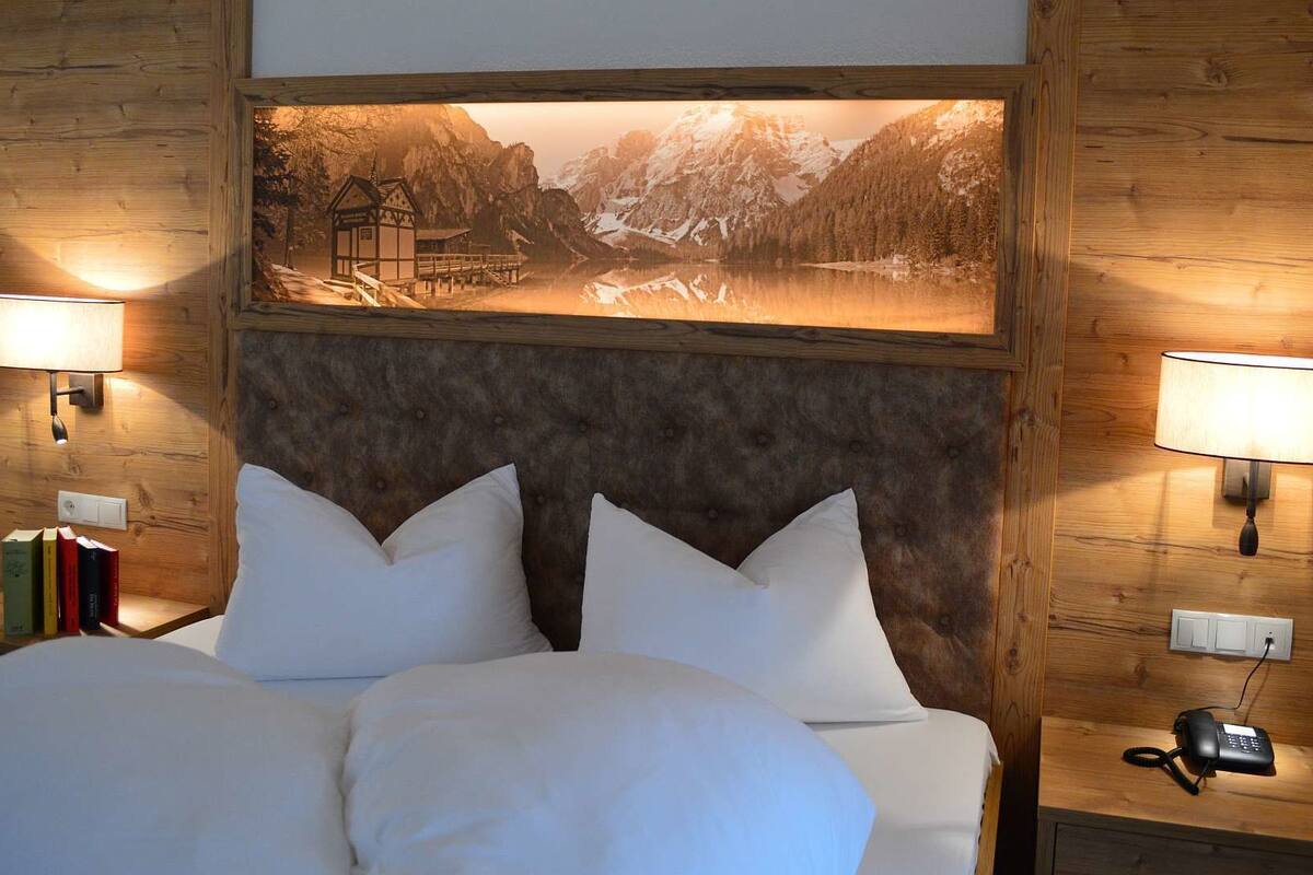 A hotel room with a bed, two bedside lamps, a telephone, and a framed picture of a mountainous landscape.