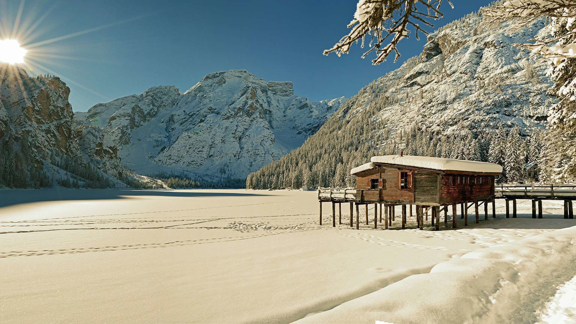 Una cabaña de madera sobre pilotes junto a un lago cubierto de nieve, con montañas al fondo.