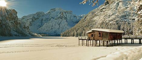 A wooden cabin on stilts by a snow-covered lake with mountains in the background.