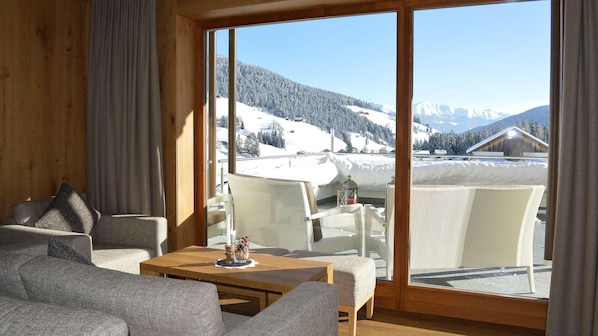 A cozy room with a large window offering a snowy mountain view. There is a sofa and a small table with a candle.