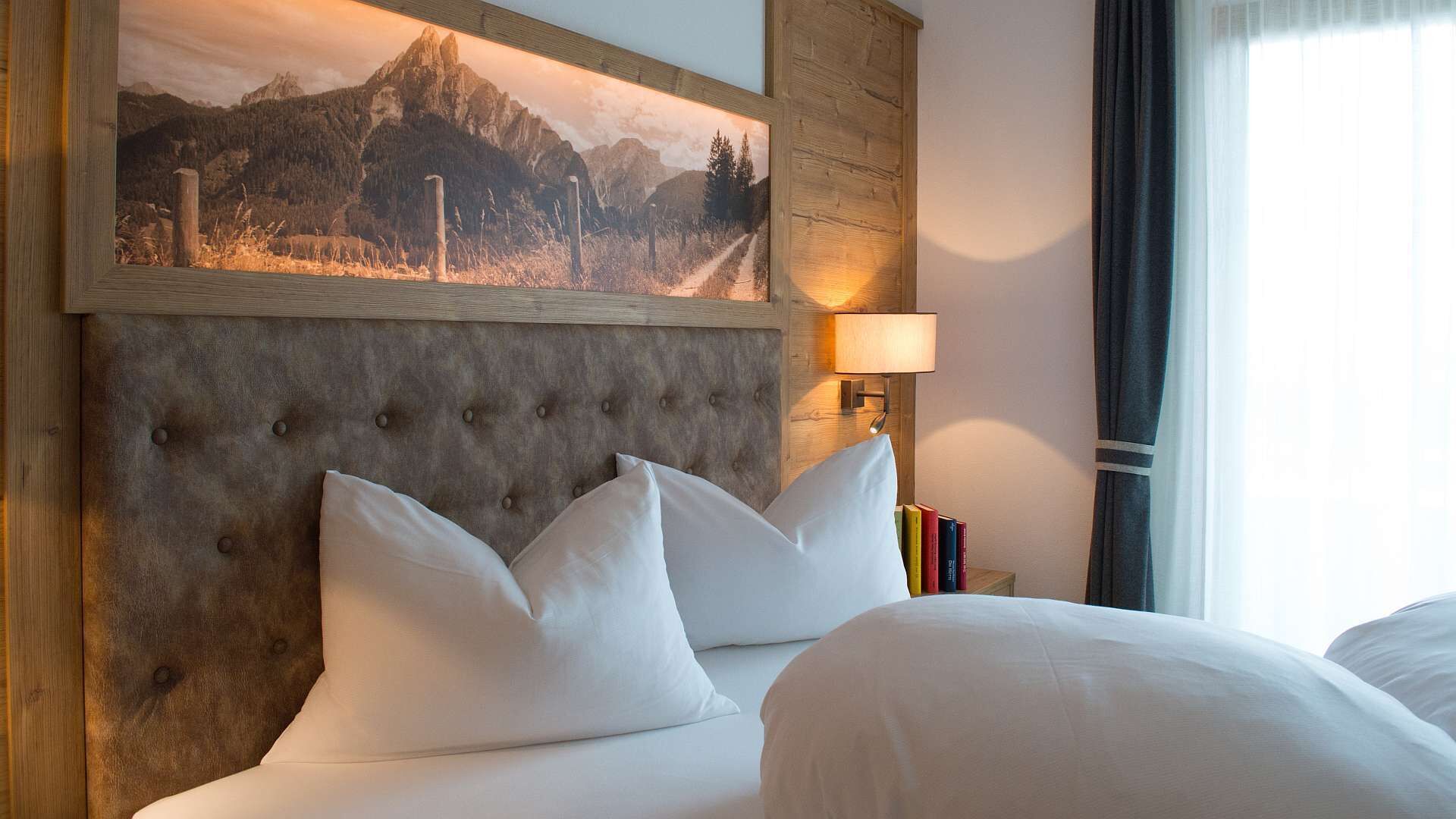 A hotel room with a large bed, a headboard with a mountain painting, a bedside lamp, and a window with curtains.