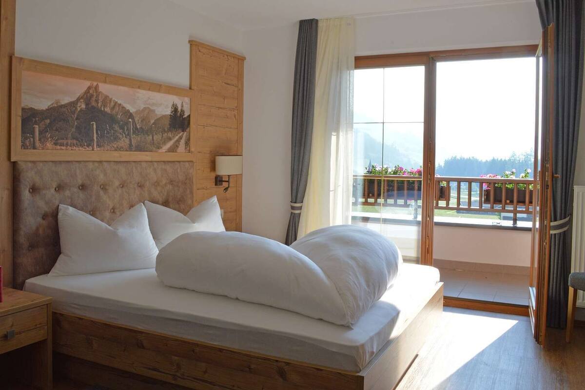 A hotel room with a wooden headboard, a bed with white linens, a nightstand with books, a wall-mounted lamp, and a balcony with a view of the mountains.