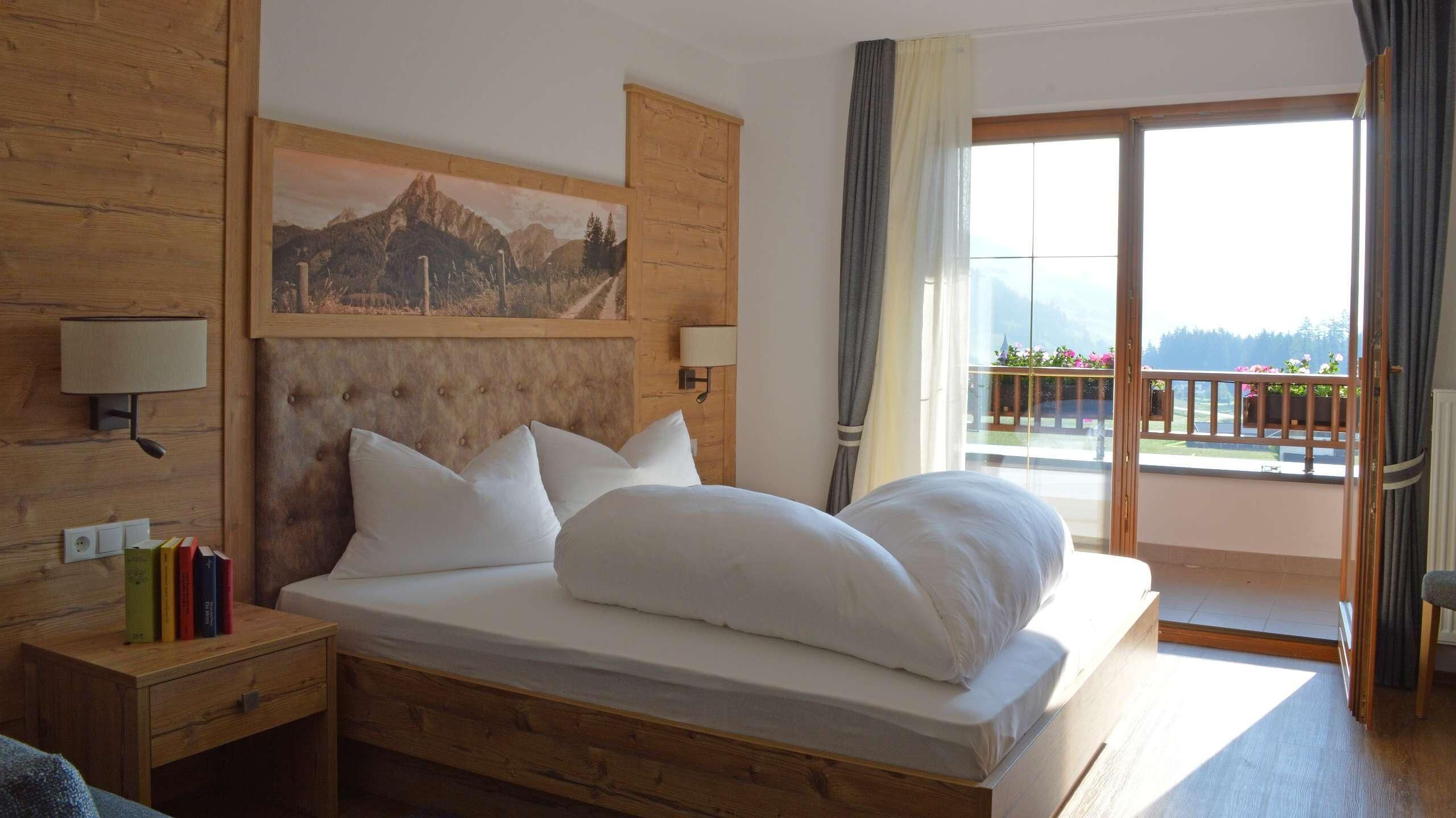 A hotel room with a large bed, a wooden headboard, a painting above the bed, a nightstand with books, a lamp, and a balcony with a view.