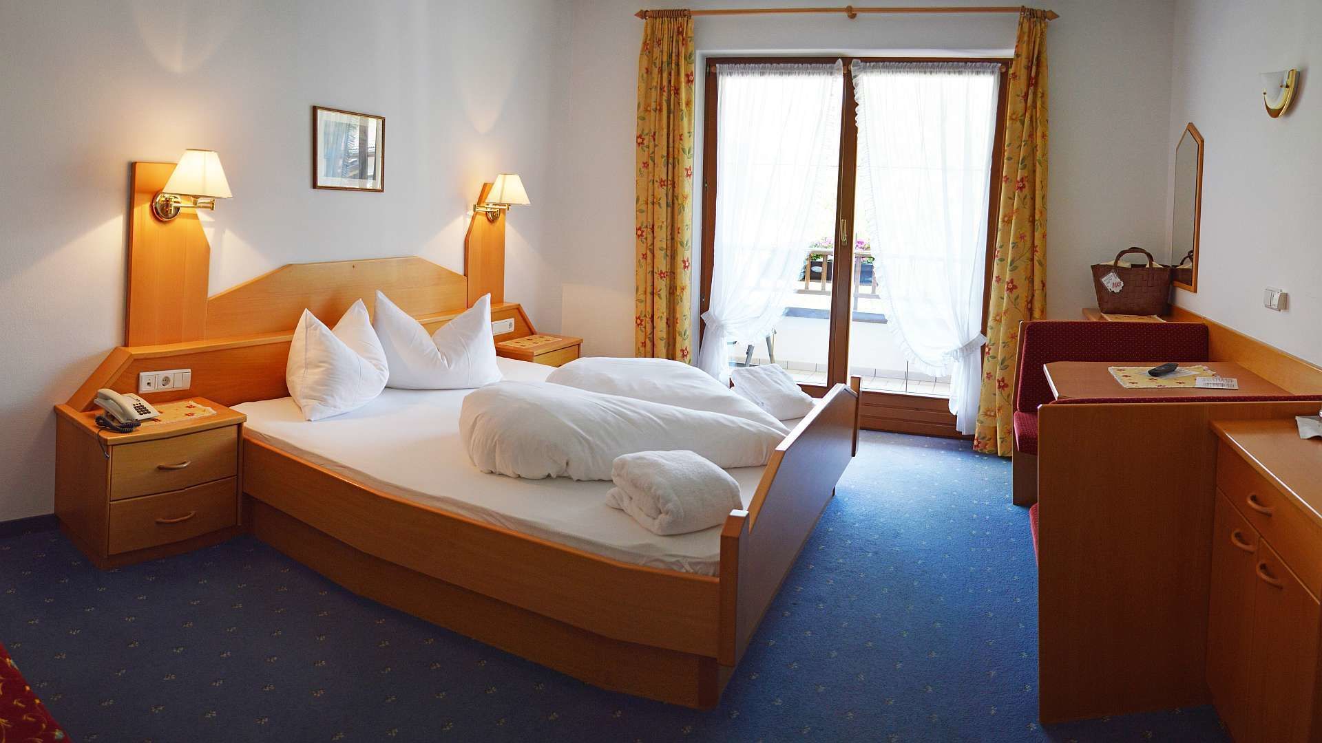 A hotel room with a bed, bedside lamps, a desk, a chair, a dresser, and a window with curtains.