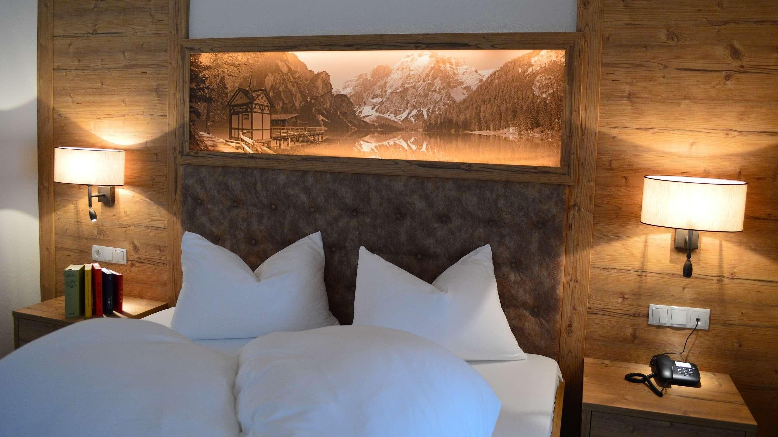 A bedroom with a large bed, two bedside tables, a mounted picture of a mountain landscape, and two wall-mounted lamps.