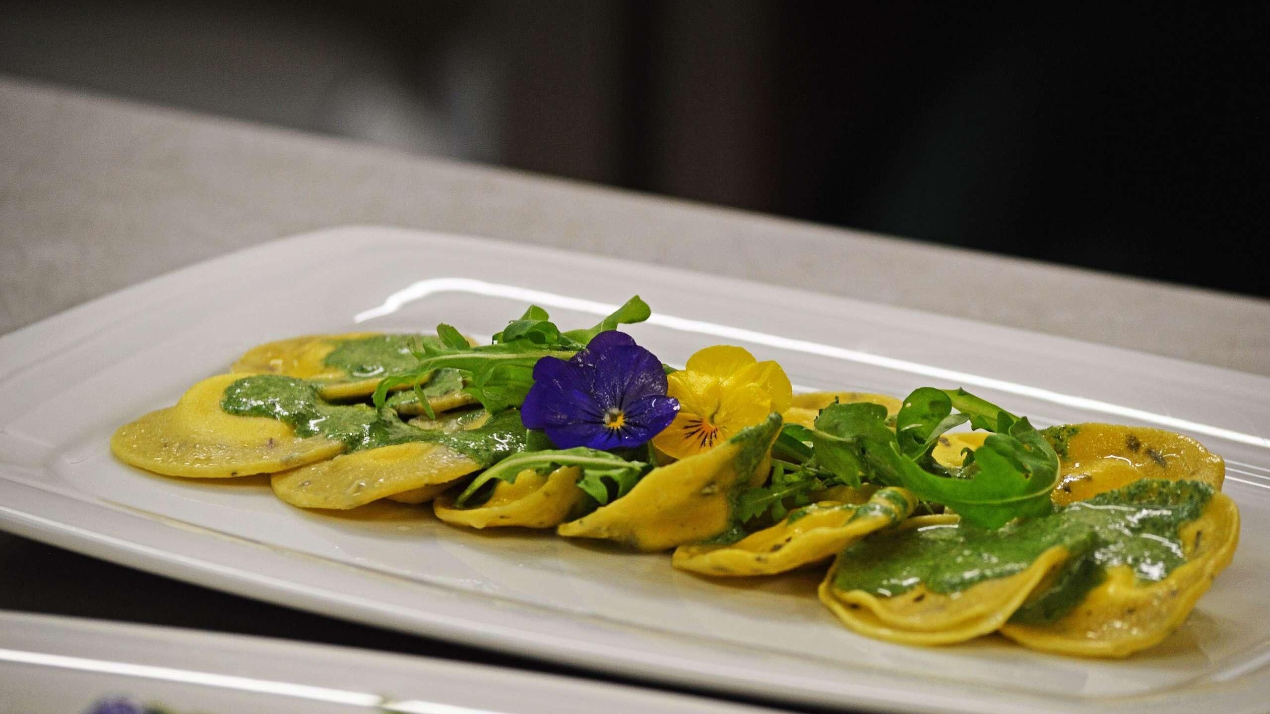 A plate of ravioli with a green sauce and garnished with a purple flower and green leaves.