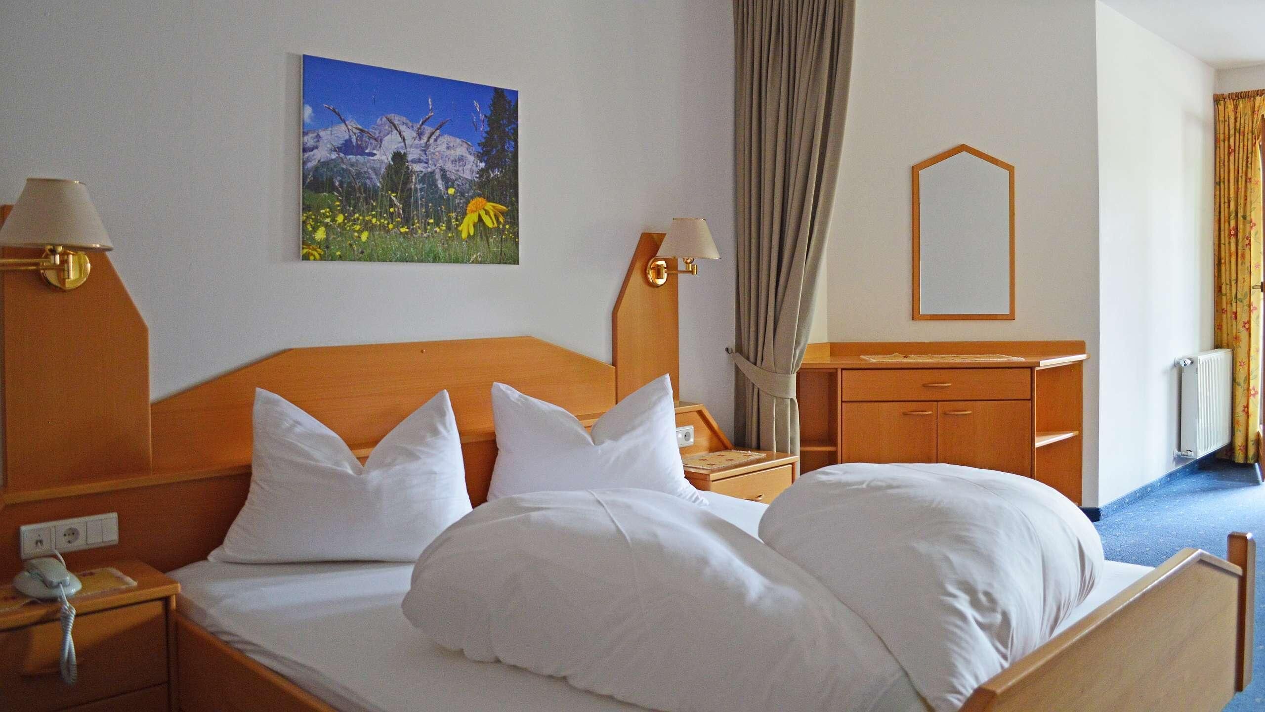 Une chambre d’hôtel avec un lit en bois, du linge de lit blanc, un tableau représentant un paysage de montagne, un miroir, une table de chevet avec une lampe et une commode.