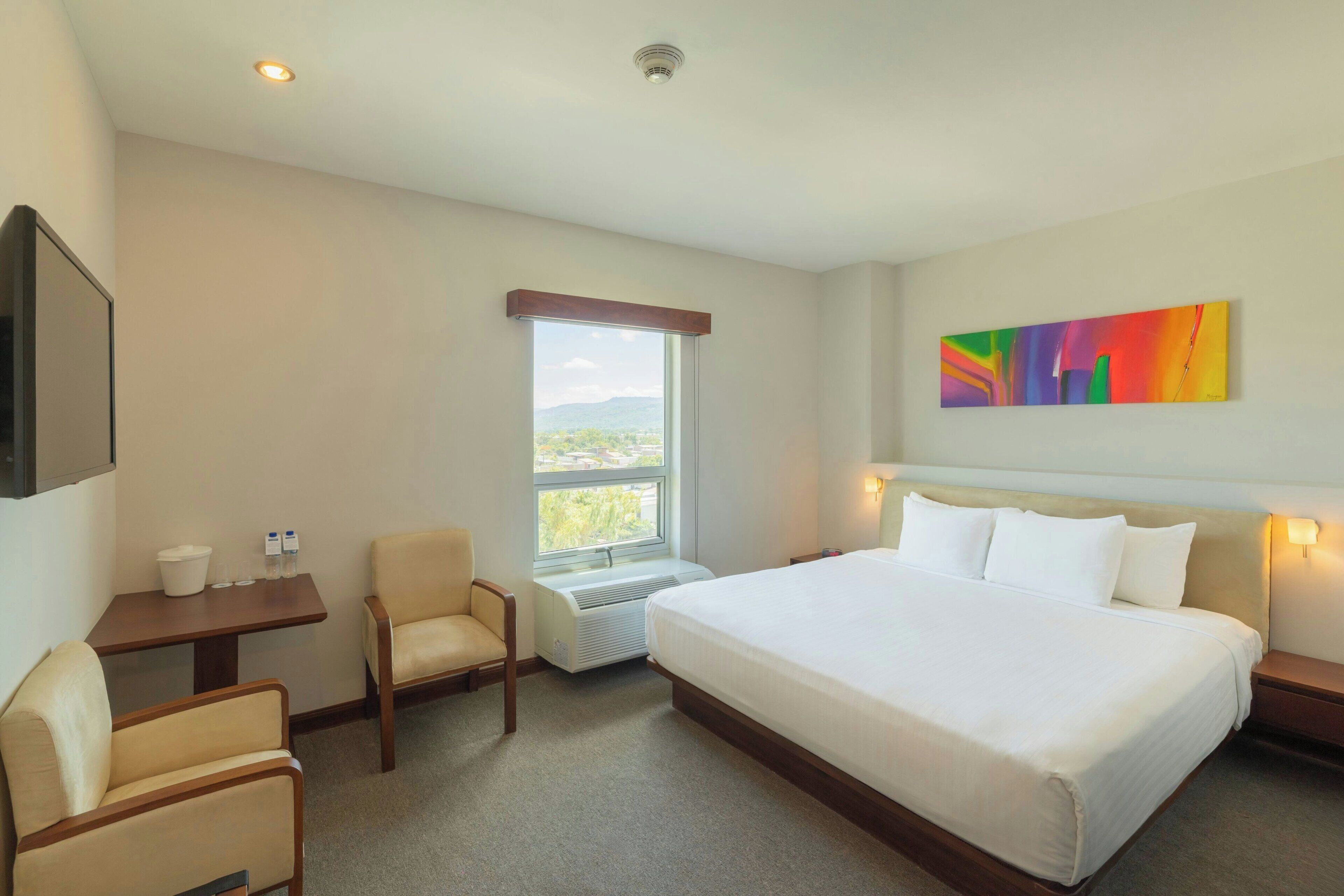Quarto de hotel com cama, duas cadeiras, escrivaninha, janela com vista, um quadro na parede e televisão montada.