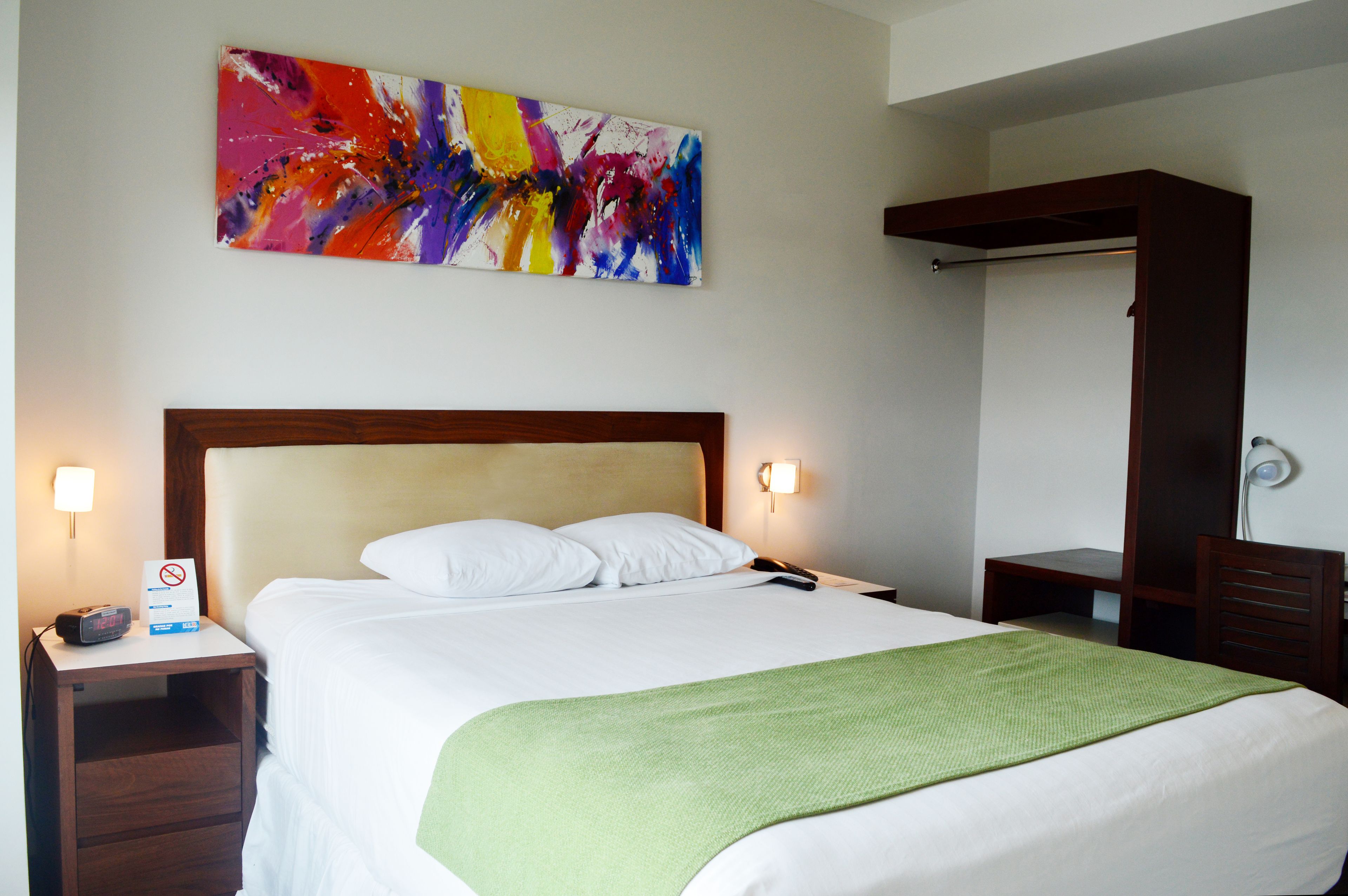 A hotel room with a large bed, a green throw blanket, a nightstand with a clock, a wall-mounted abstract painting, and a wooden wardrobe.