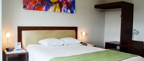 Una habitación de hotel con una cama grande, una manta verde, una mesita de noche con un reloj, un cuadro abstracto en la pared y un armario de madera.