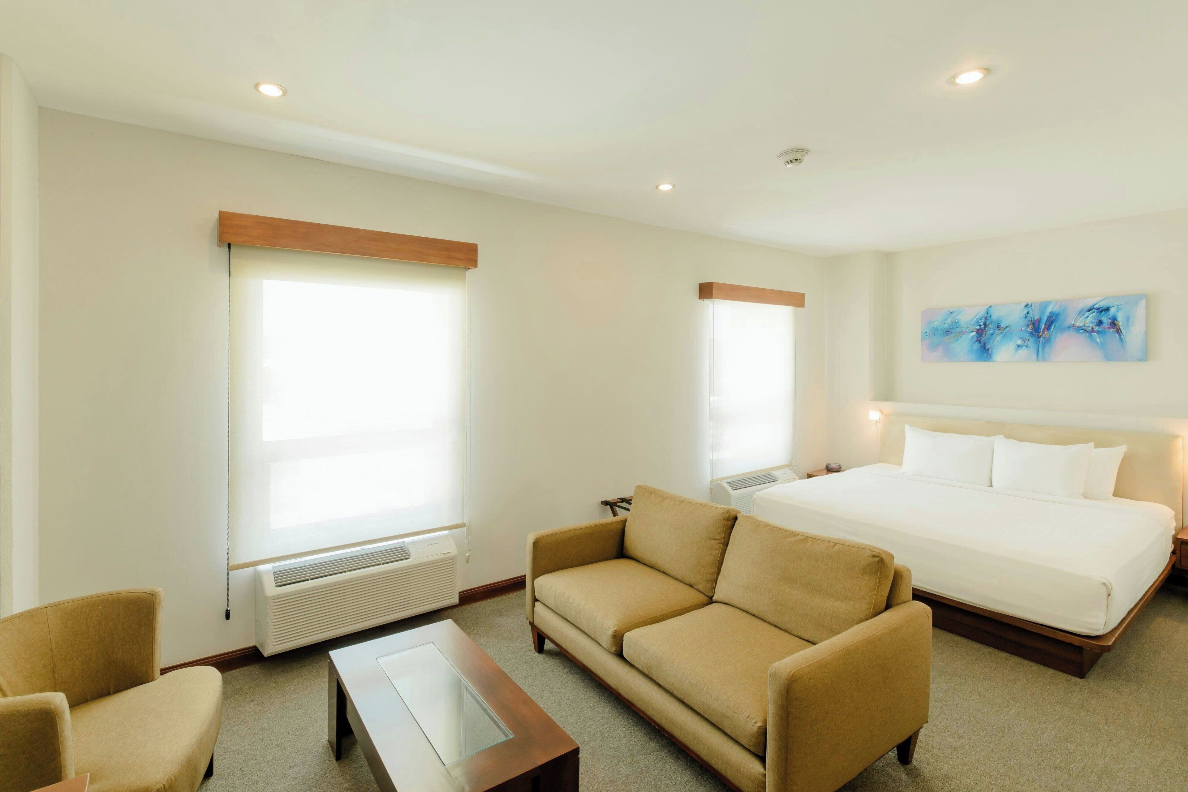 Une chambre d’hôtel comprenant un lit, un canapé, une table basse et deux fenêtres équipées de stores.