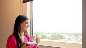 Una persona sosteniendo una taza y un plato junto a una ventana con vistas a edificios y árboles.