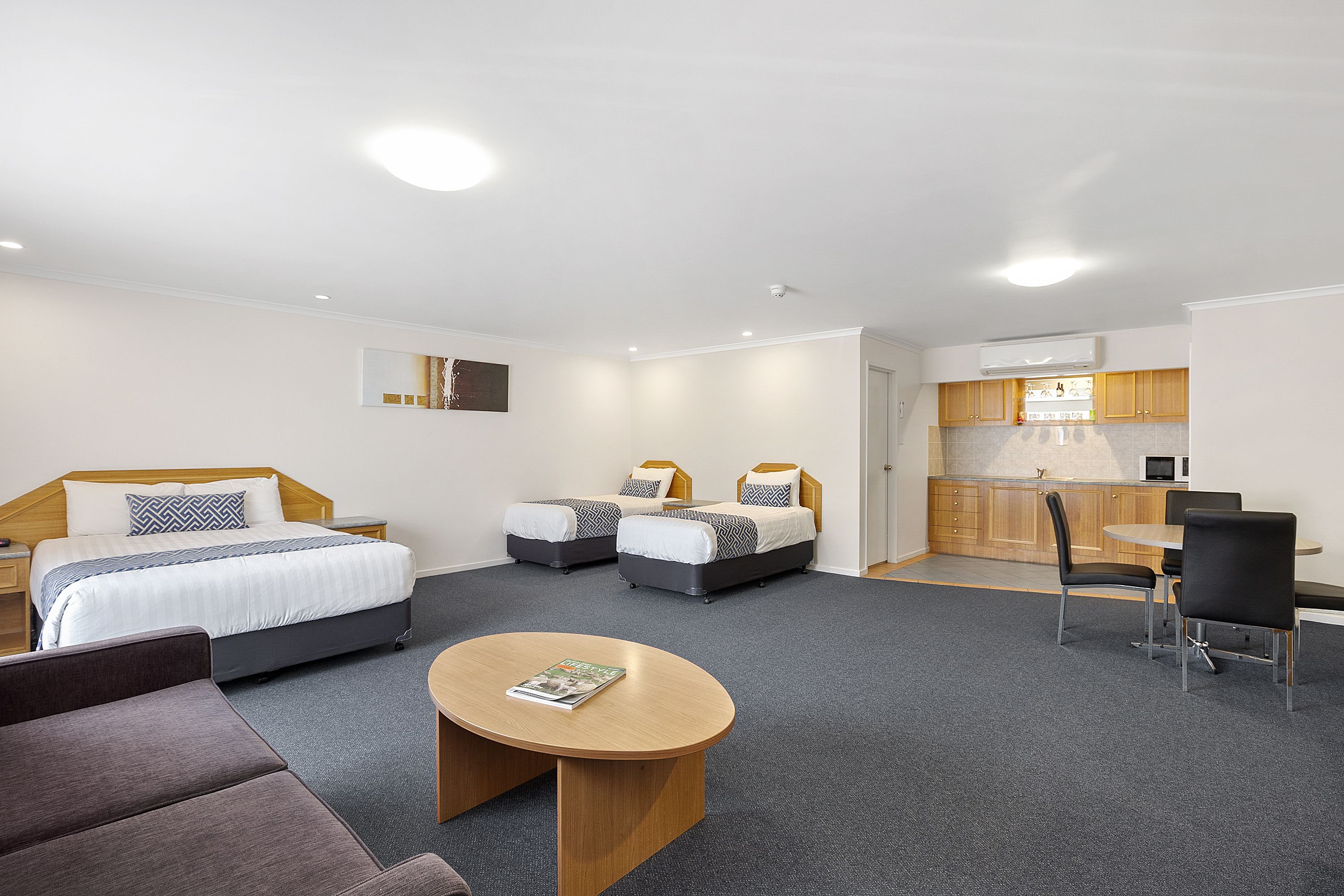 Una habitación de hotel moderna con una cama grande, dos camas más pequeñas, un comedor con mesa y sillas, y una kitchenette.