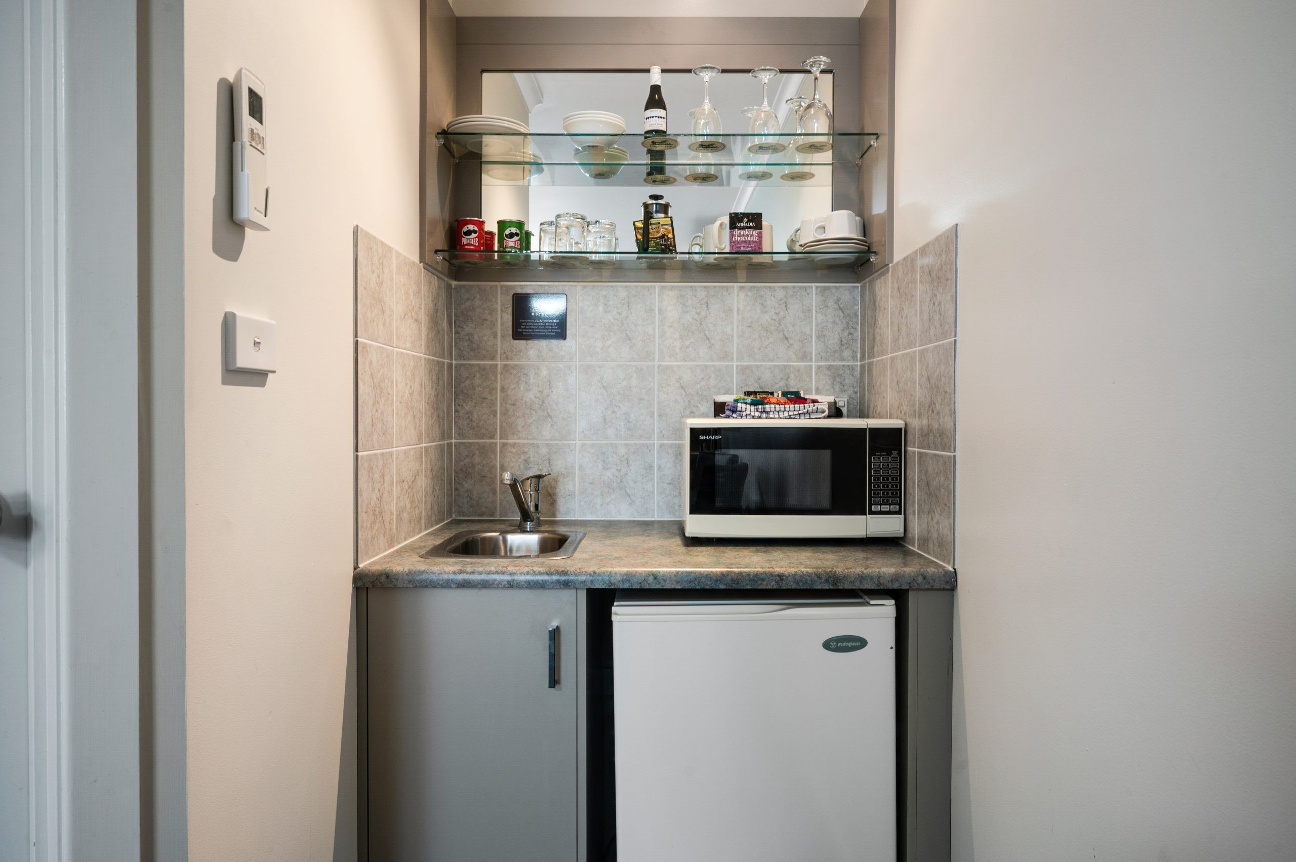 A compact kitchen with a built-in microwave, a small refrigerator, and a glass shelf above the sink holding various items.
