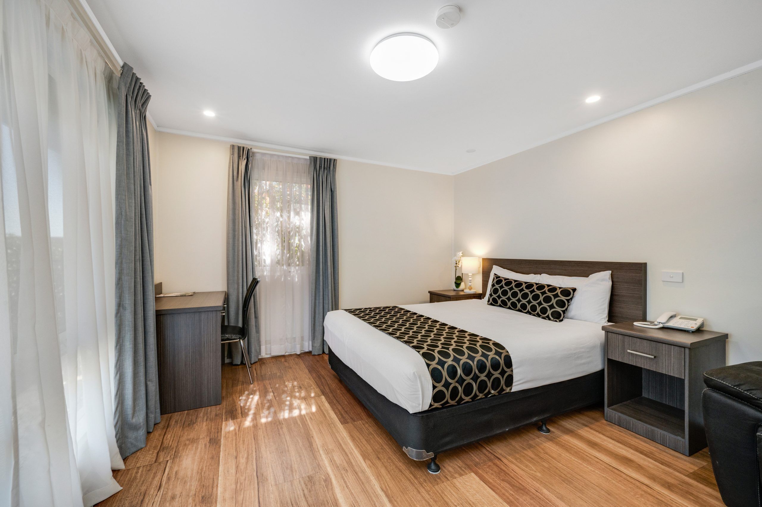 A hotel room with a bed, bedside table, lamp, and a desk with a chair.