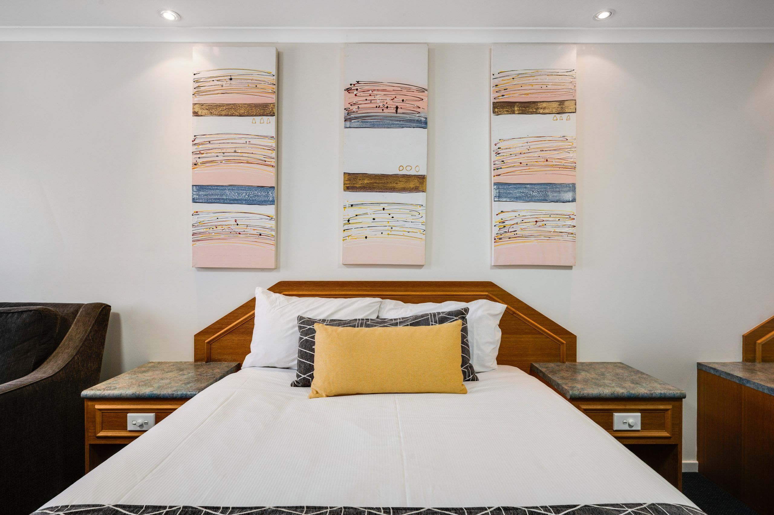 Habitación de hotel con una cama, dos mesitas de noche, un cabecero de madera y tres cuadros abstractos en la pared.