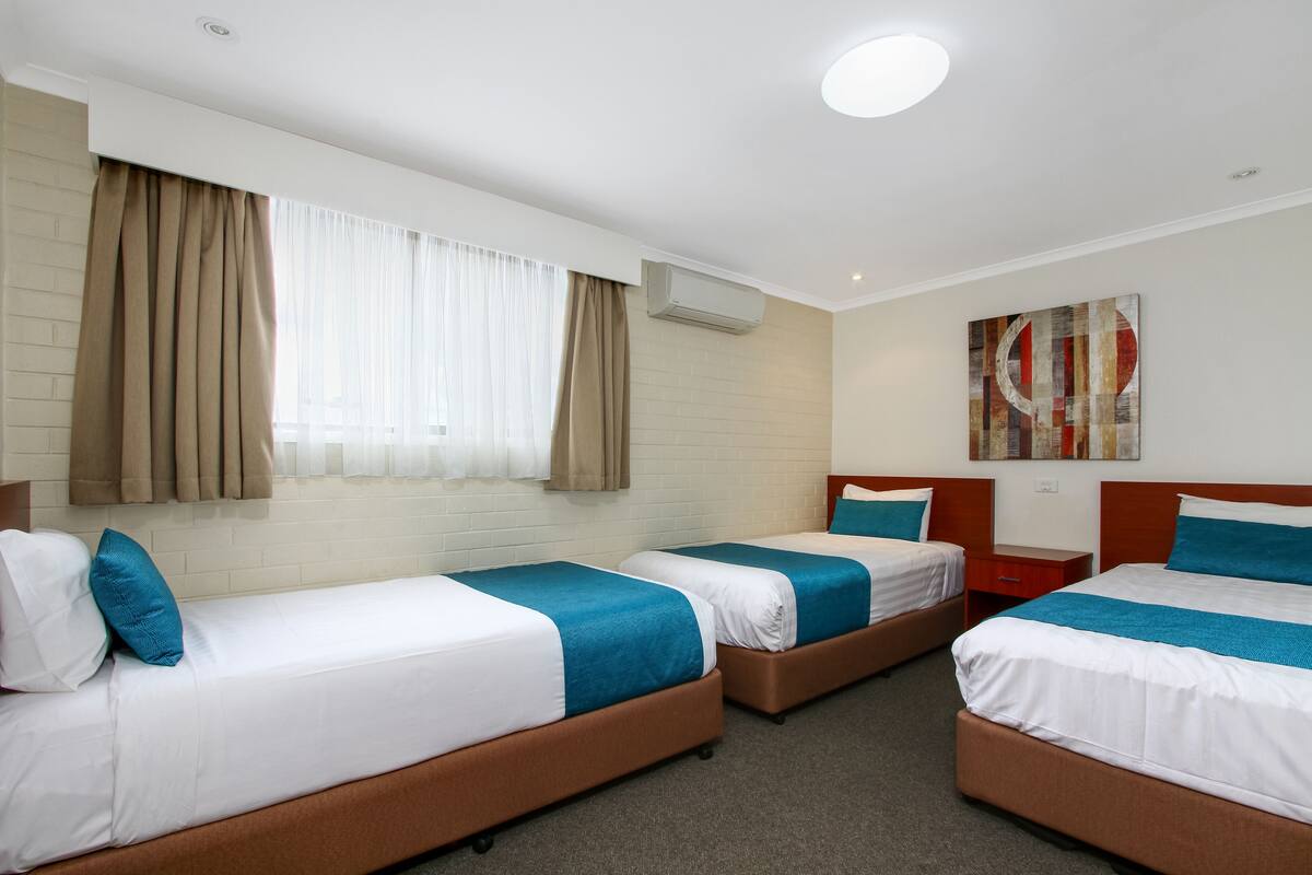 Ein Hotelzimmer mit vier nebeneinander stehenden Betten, einem Fenster mit Vorhängen, einer Klimaanlage und einem Wandbild.