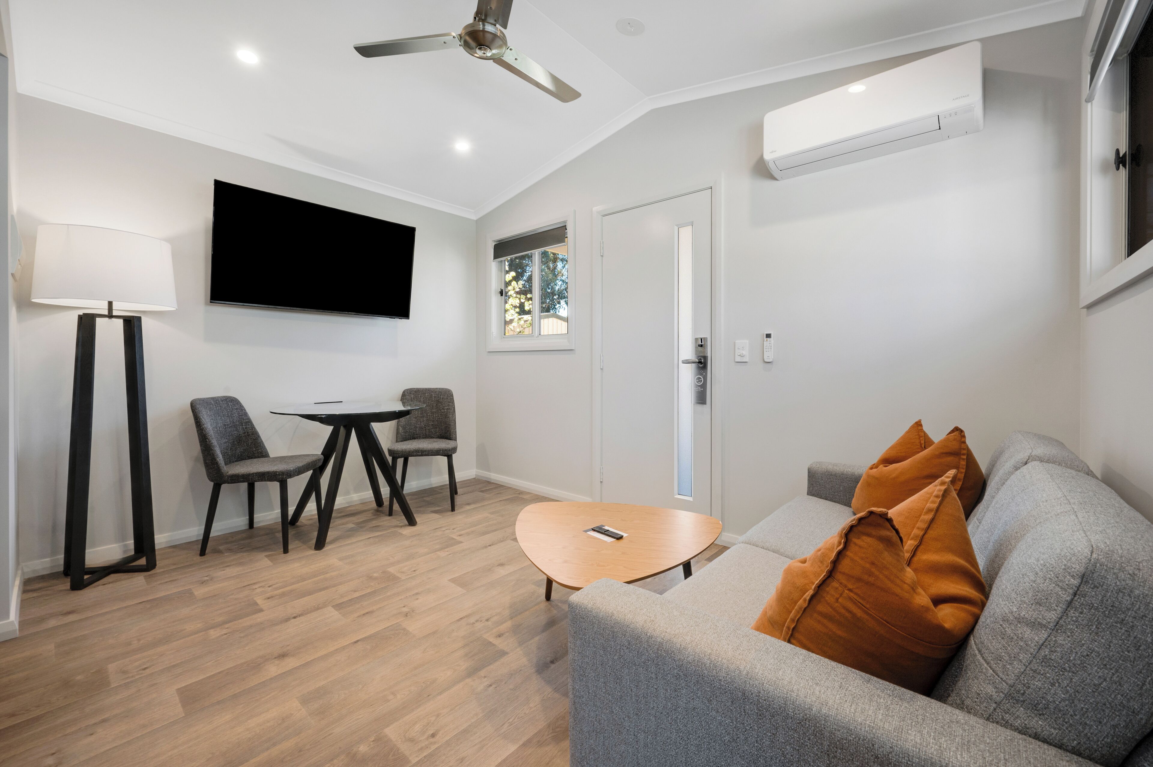 A modern living room with a grey sofa, a small round table, and two chairs. There is a flat-screen TV mounted on the wall, a ceiling fan, and an air conditioning unit.