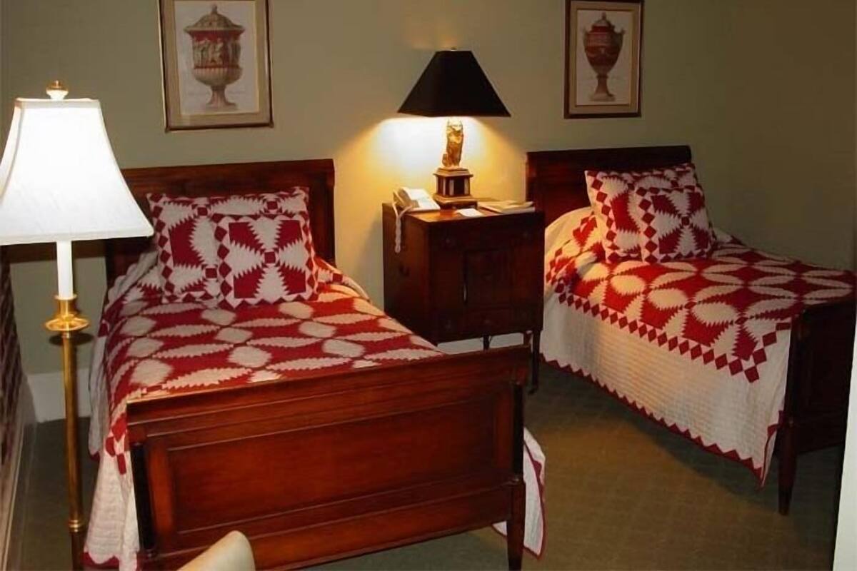 A room with two wooden beds, each with a red and white patterned bedspread. There is a nightstand with a lamp between the beds, and two framed pictures on the wall.