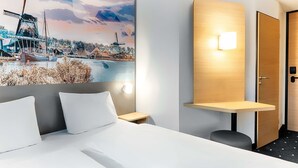 Une chambre d’hôtel comprenant un lit, une table de chevet, une lampe fixée au mur, une porte et une grande fresque représentant un moulin à vent et un paysage aquatique.