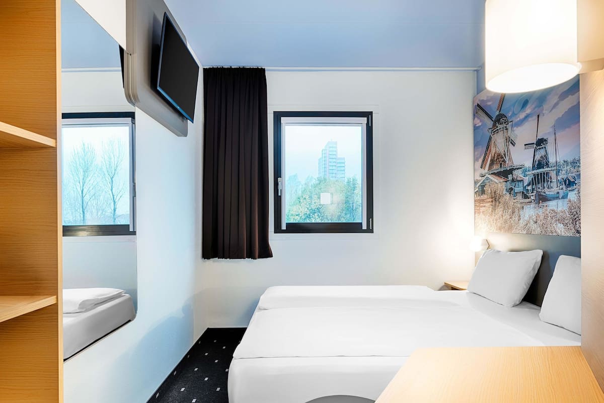 Een hotelkamer met een bed, een aan de muur bevestigde televisie, een raam met uitzicht op gebouwen en een schilderij van windmolens.