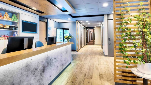 Un espace d’accueil moderne comprenant un comptoir de réception en bois, un réfrigérateur et une plante.