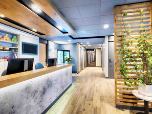 A modern reception area with a wooden reception desk, a refrigerator, and a plant. - B&B Hotel Amsterdam-Zaandam (Zaandam)