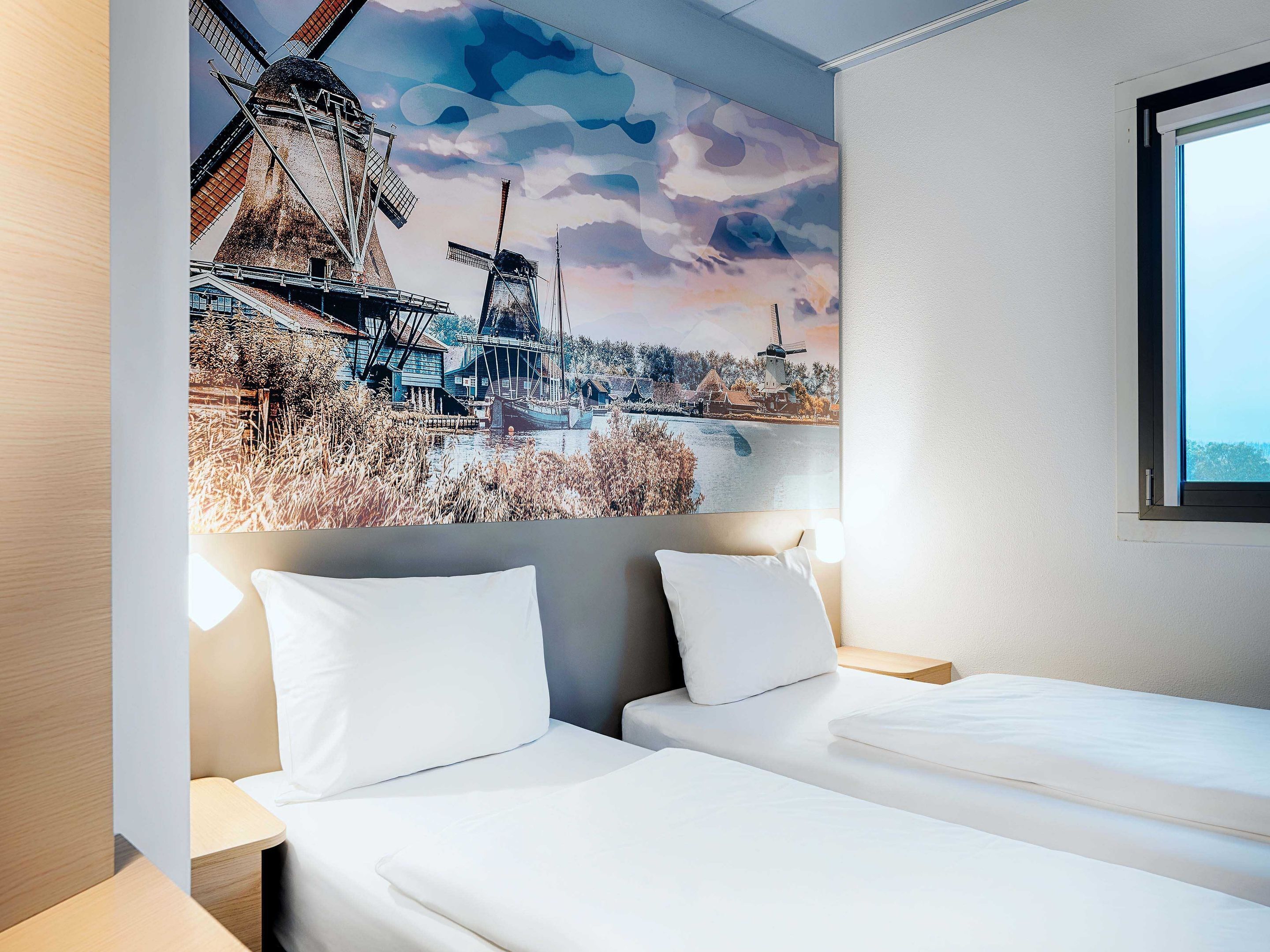 A hotel room with a bed, two pillows, a nightstand, a lamp, and a large painting of a windmill and water scene on the wall.