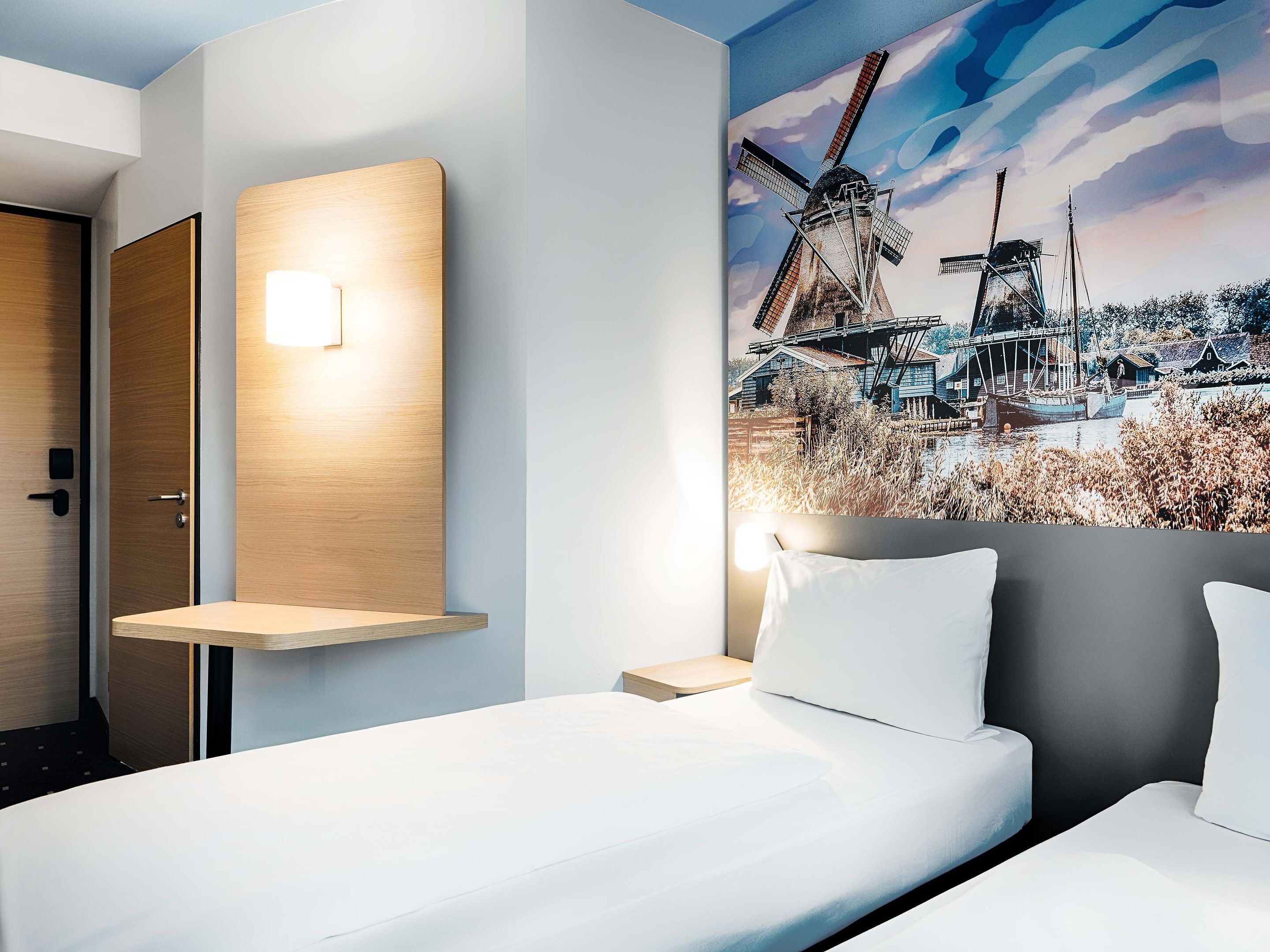 Una camera d'albergo con un letto, comodini, una lampada a parete e un grande quadro raffigurante un mulino a vento.
