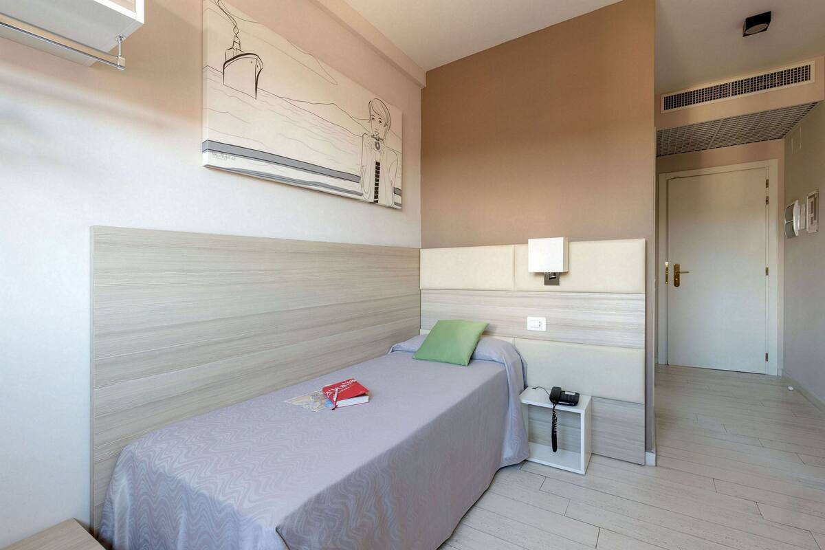 A single bed with a grey bedspread, a green pillow, and a bedside table with a phone.