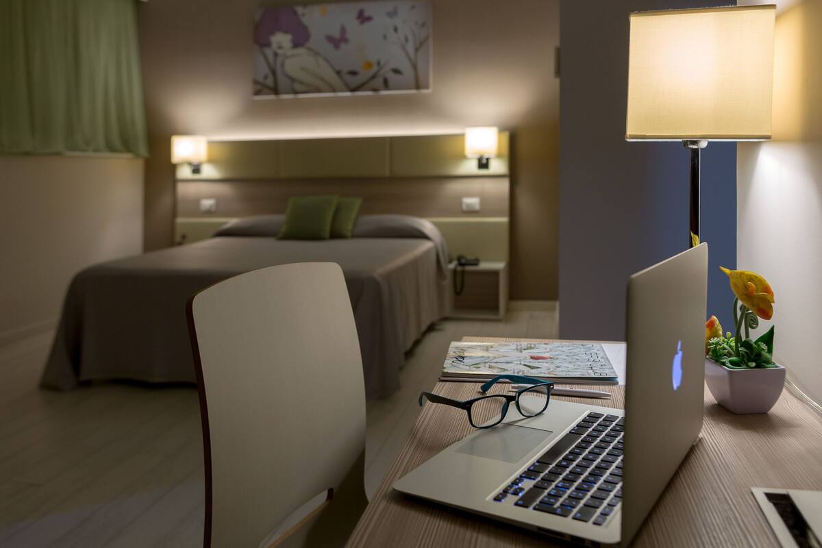 A hotel room with a bed, a desk with a laptop, glasses, a lamp, and a small plant.