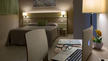 A hotel room with a bed, a desk with a laptop, glasses, a lamp, and a small plant.