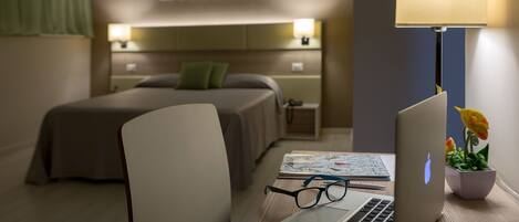 A hotel room with a bed, a desk with a laptop, glasses, a lamp, and a small plant.