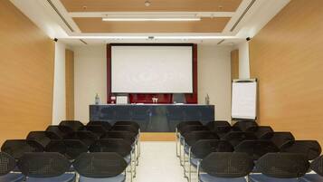 A conference room with rows of chairs facing a projection screen, a whiteboard, and a small table with a bottle and a glass.