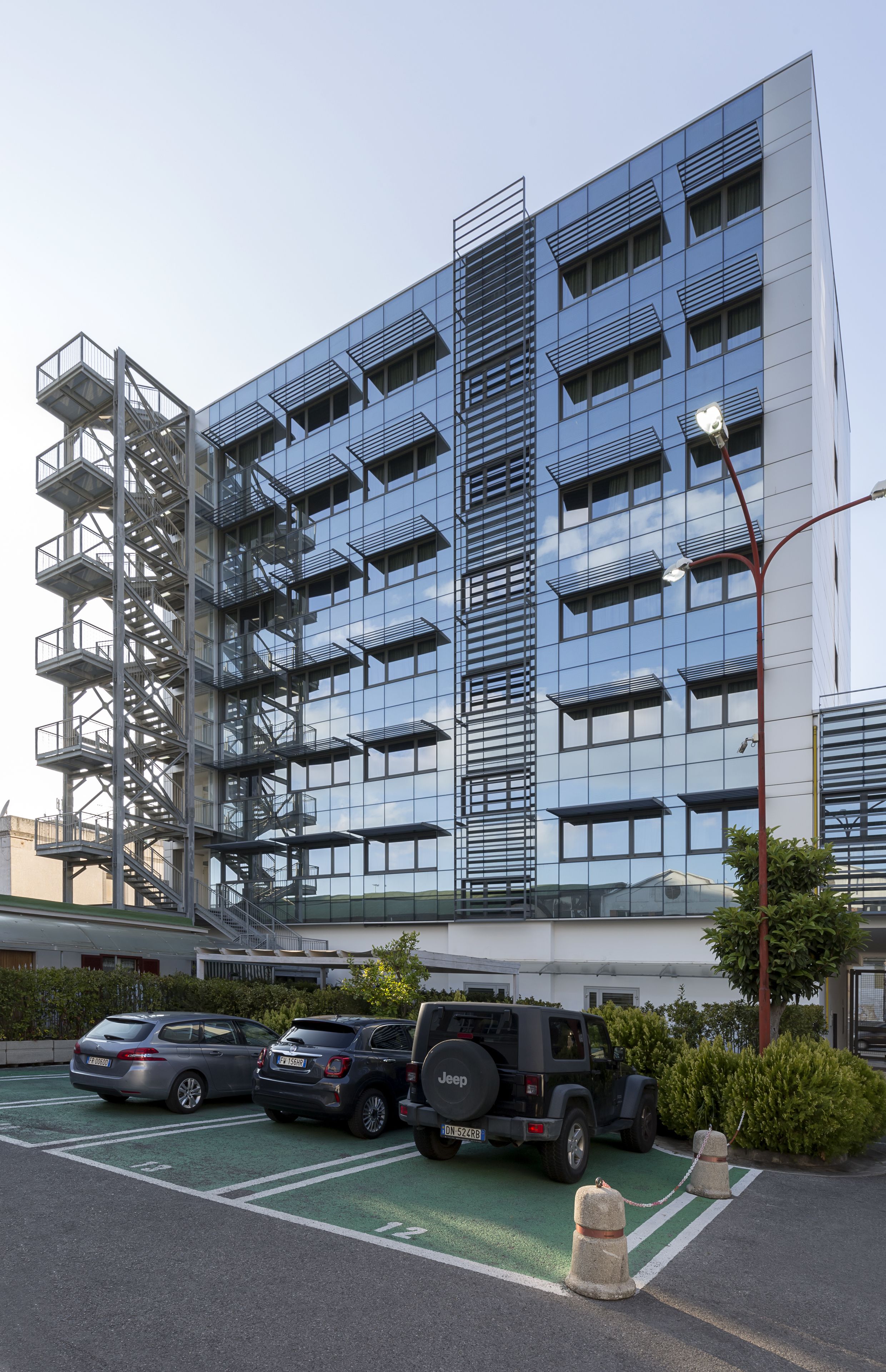 Un edificio moderno con fachada de vidrio, una escalera de incendios y un estacionamiento con coches y un Jeep.