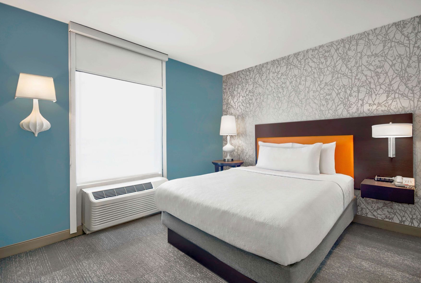 A hotel room with a bed, two bedside tables, a window with blinds, a wall-mounted lamp, and a patterned wallpaper.