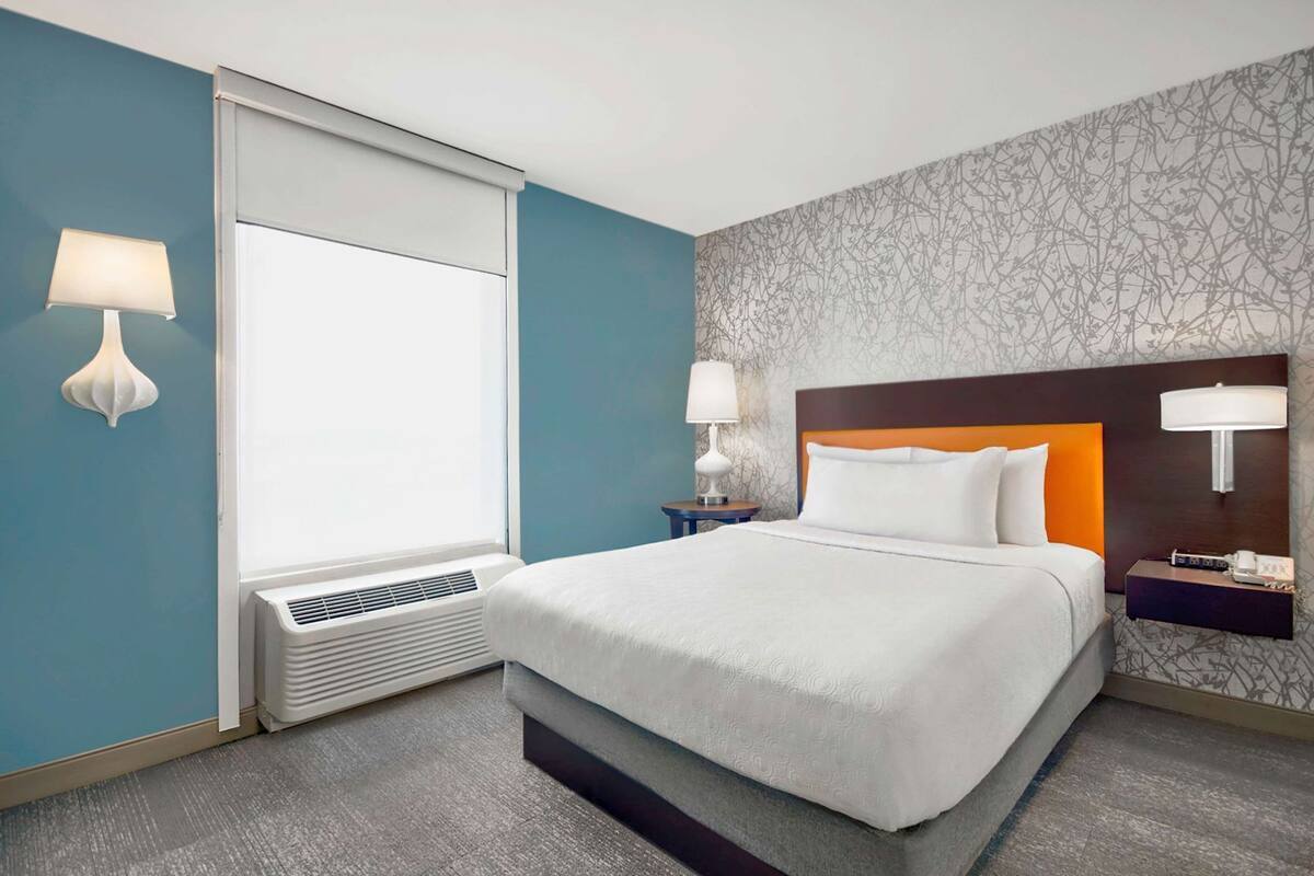 A hotel room with a bed, two bedside tables, a window with blinds, a wall-mounted lamp, and a patterned wallpaper.