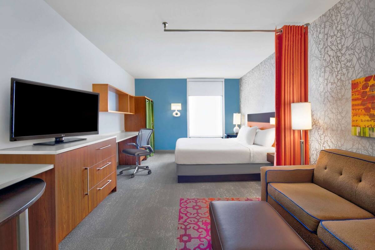 A hotel room with a bed, a TV, a desk with drawers, a chair, a sofa, and a window.